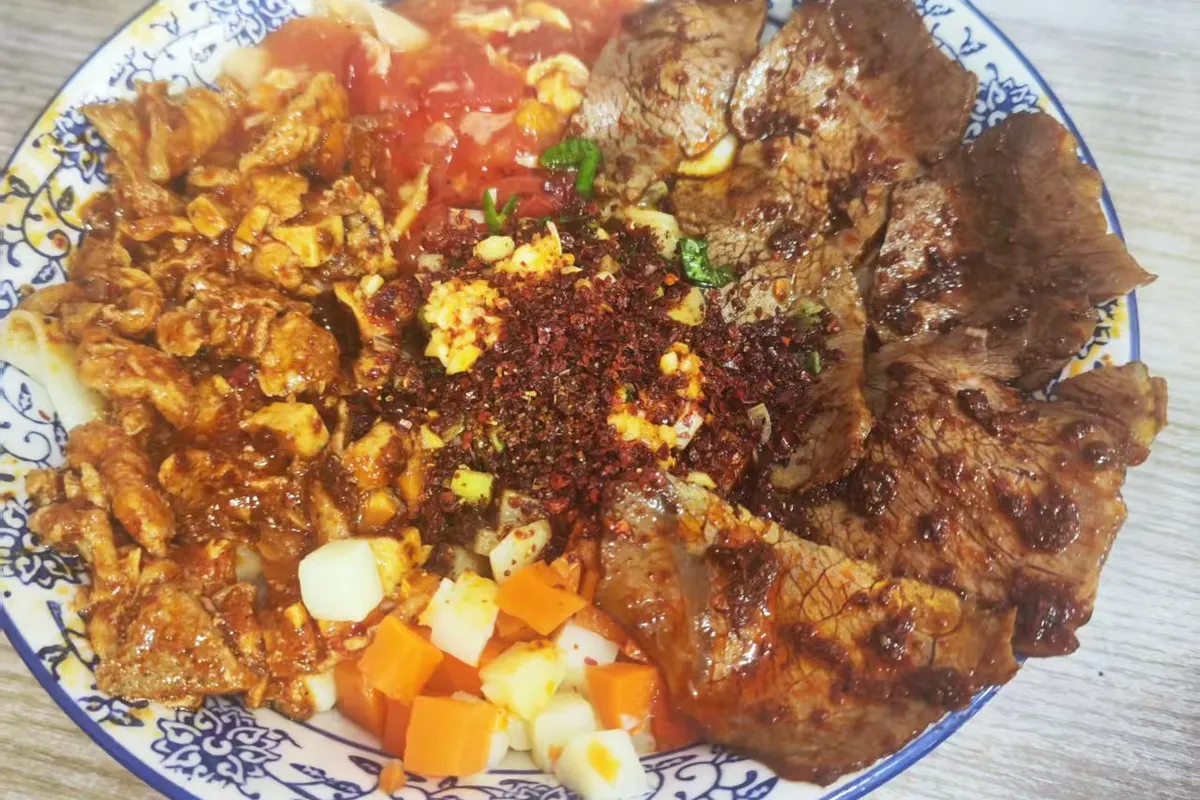 A loaded bowl of biangbiang noodles on a blue-and-white plate — belt noodles under braised beef slices, tomato-and-egg, minced meat, diced potato and carrot, with raw garlic and chili flakes heaped in the center