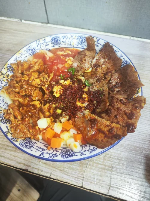 A loaded bowl of biangbiang noodles on a blue-and-white plate — belt noodles under braised beef slices, tomato-and-egg, minced meat, diced potato and carrot, with raw garlic and chili flakes heaped in the center