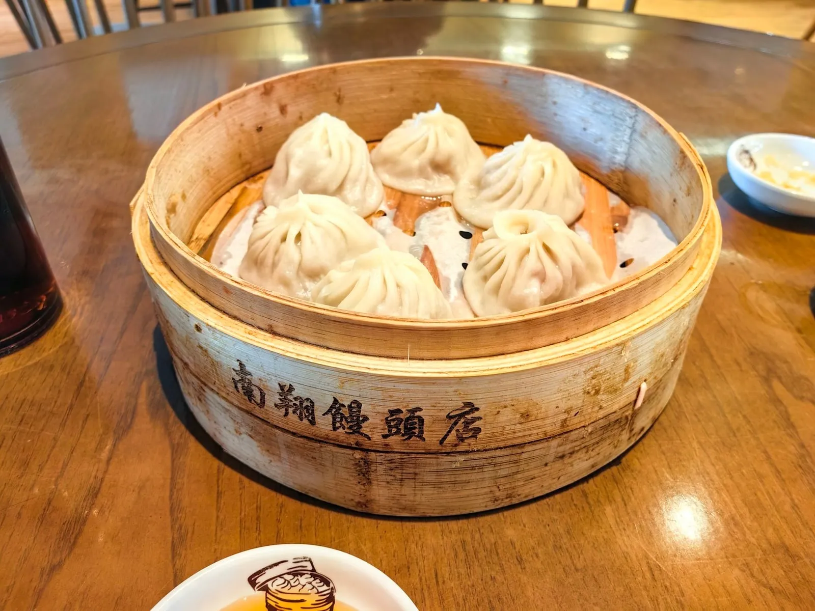 Four pleated xiaolongbao in a bamboo steamer marked Nanxiang on a wooden table