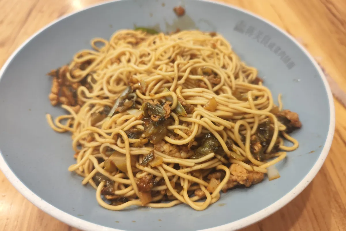 A bowl of xuecai noodles tossed dry — pickled mustard greens and shredded pork mixed through the noodles — in a gray-blue ceramic bowl on a wooden table