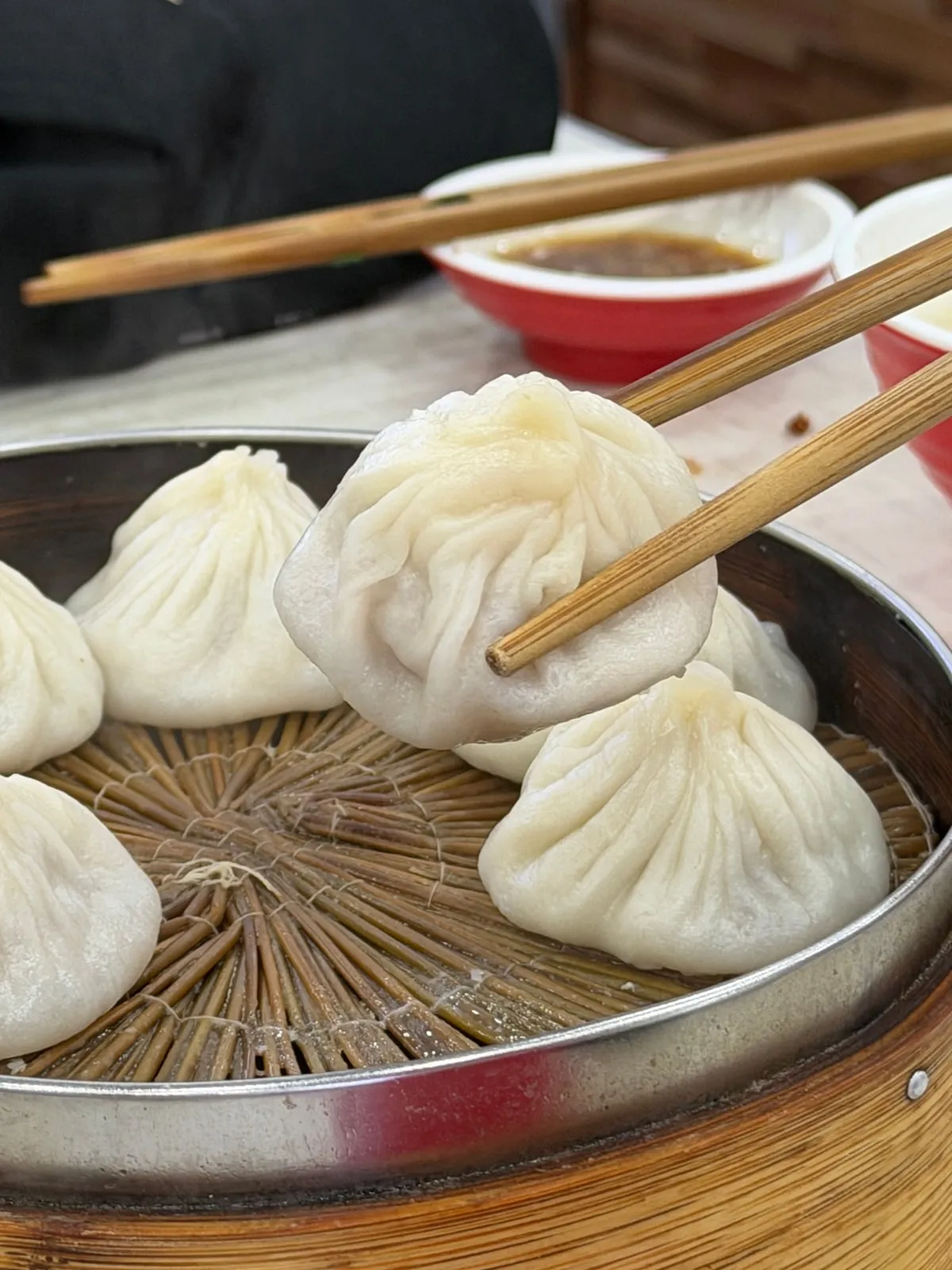 Chopsticks gently lifting a thin-skinned soup dumpling above a bamboo steamer at Shanshan
